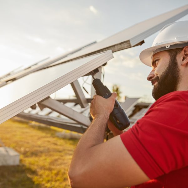 Side view of bearded male technician in hardhat and uniform with screwdriver installing solar panel while working at station producing renewable power
